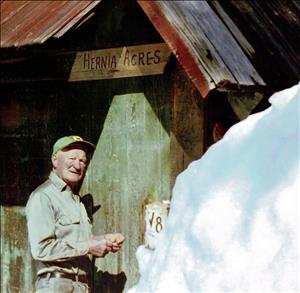 White man stands in front of a snowbank and a wooden building next to a sign that says Hernia Acres
